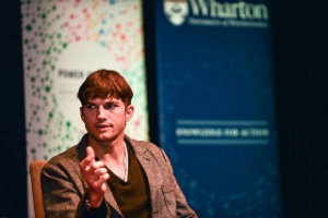 Close-up of Ashton Kutcher, talking in front of a Wharton banner