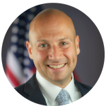 Elad Roisman in front of the American flag, wearing a black blazer and white collared shirt and tie