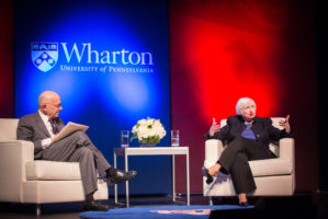 Janet Yellen and Jeremy Siegel sit on stage, in conversation, with large red and blue panels behind them
