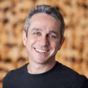 A headshot of a smiling person with short gray hair, wearing a black shirt, against a blurred, warm-toned background.