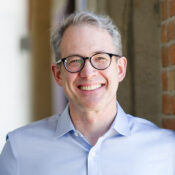 A headshot of a person wearing glasses and a light blue shirt, smiling, with short gray hair, standing near a brick wall.