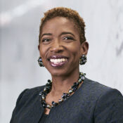 Headshot of a person smiling, wearing earrings and a necklace, dressed in a dark textured jacket with a blurred gray background.