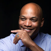 A smiling person in a light blue shirt and tie, resting their chin on their hand against a dark background.