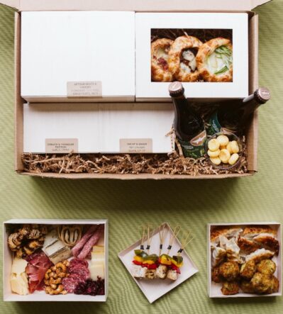 A gourmet meal box containing assorted foods: artisan meats and cheeses, small pies, skewers, dumplings, with two bottled beverages.