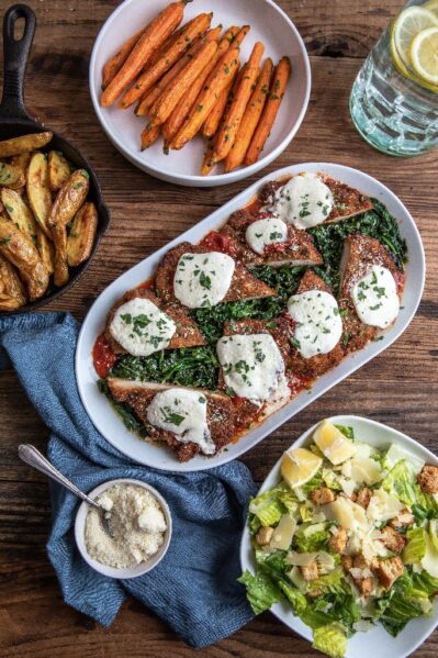 A wooden table spread with a meal: a platter of chicken breasts topped with sauce and mozzarella, roasted carrot sticks, roasted potato wedges in a skillet, Caesar salad, parmesan in a small dish, and a glass of lemon water.