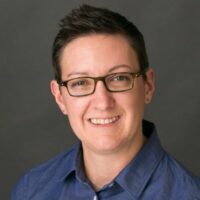 Headshot of a person with short dark hair, glasses, and a blue collared shirt, smiling against a gray background.