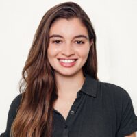 A headshot of a person with long brown hair smiling, wearing a dark button-up shirt, set against a plain background.