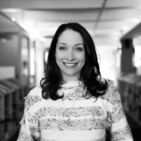 Headshot of a person with long hair, smiling and wearing a patterned sweater. The setting appears to be indoors with a blurred hallway background.