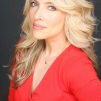 Headshot of a person with long, wavy blonde hair wearing a bright red top and a delicate necklace, set against a dark background.