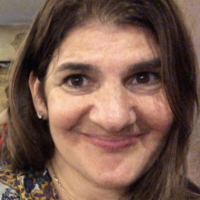 Headshot of a person with long brown hair, smiling. Wearing a patterned top and a necklace.