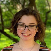 A headshot of a person with short brown hair and glasses, smiling outdoors with a background of green trees and foliage.