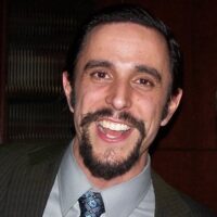 A person with dark hair and a goatee is smiling broadly, dressed in a suit and tie.