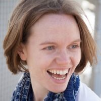 A person smiling broadly, with short brown hair, wearing a blue scarf and a light-colored top.