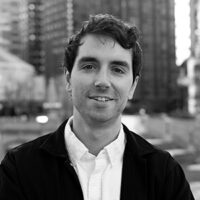 A grayscale headshot of a person with short hair, wearing a collared shirt and jacket, in an urban setting with blurred buildings in the background.