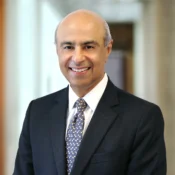 Headshot of a person wearing a dark suit, light shirt, and patterned tie, smiling in a professional setting.